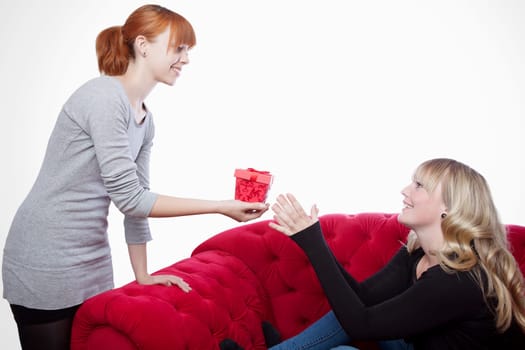 young beautiful blond and red haired girls on red sofa have a present box in front of white background