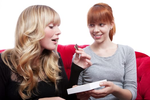 young beautiful blond and red haired girls eating chocolate on red sofa in front of white background