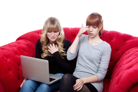 young beautiful blond and red haired girls on red sofa are shocked about news on laptop in front of white background