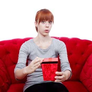 young beautiful red haired girl opens gift box for you on red sofa in front of white background