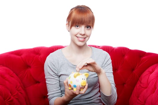 young beautiful red haired girl put coin in piggybank on red sofa in front of white background