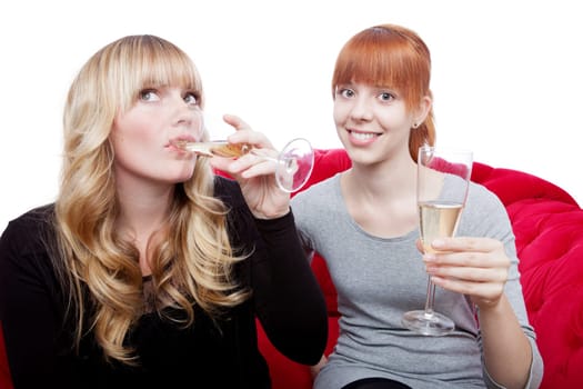 young beautiful blond and red haired girls drink champagne and cheer on red sofa in front of white background
