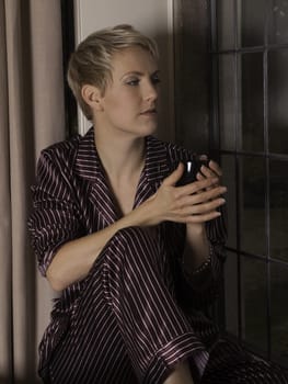Beautiful blond girl by window drinking a mug of tea or coffee.