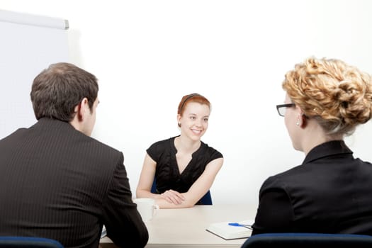 Young woman being interviewed for a job smiling as she answers questions from the two corporate interviewers, a man and woman, who are sitting with their backs to the camera