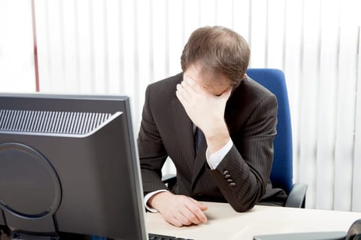 Despairing businessman sitting at his desk in his office resting his forehead in his hands as he considers the hopelessness of the situation