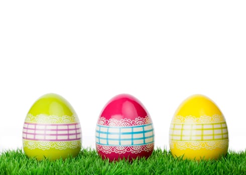Three colourful decorative Easter Eggs tied around with ornamental lacy ribbon standing on short green grass against a white background with copy space for your greeting