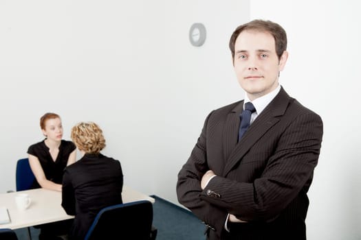 Confident handsome young male leader or manager standing with his arms folded smiling at the camera while his team go about their work in the background