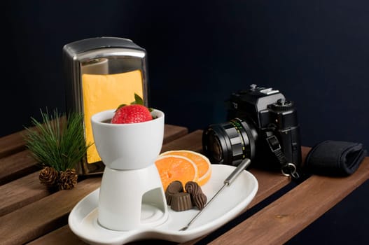 Straberry dipped into fondue with white china fondue set and camera on wooden table