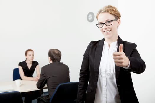 Business woman wearing glasses standing to the forefront while her colleagues work in the background giving a thumbs up of success and approval, conceptual of leadership , teamwork and authority