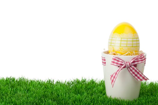 Pretty Easter gift with a decorative yellow Easter Egg inside a flower pot tied with a rustic red and white checked ribbon and bow standing on green grass against a white background with copyspace