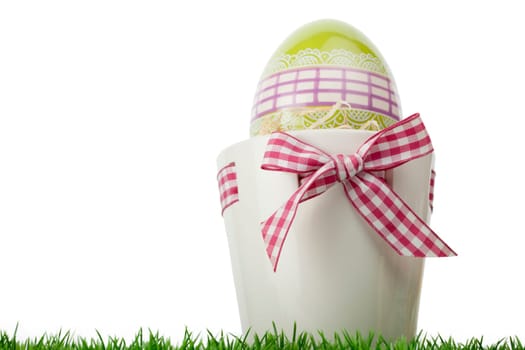 Low angle view of a decorative Easter Egg gift with a green ornamental l egg iside a flower pot tied with a fresh red and white checked ribbon and bow on grass isolated on white with copy space