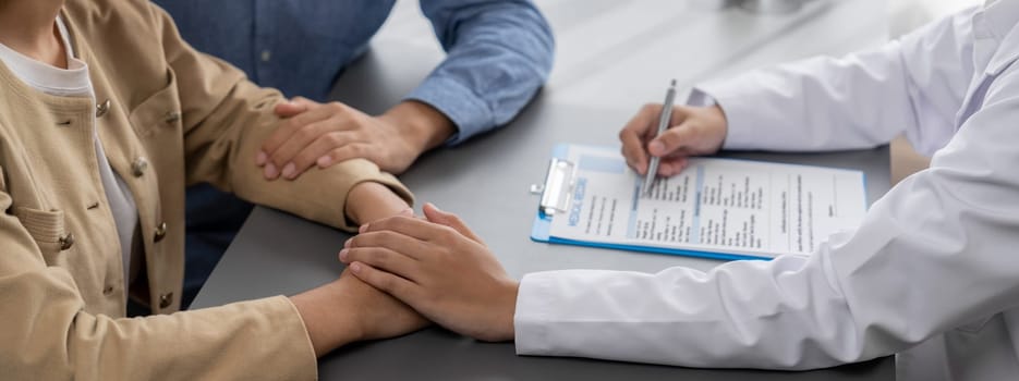 Doctor providing compassionate healthcare consultation while young couple patient holding hand, comfort each other after infertile report. Reproductive and medical fertility consulting. Neoteric