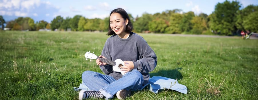 Happy brunette asian girl, student playing ukulele sitting on grass in park, relaxing, singing song, lifestyle concept.