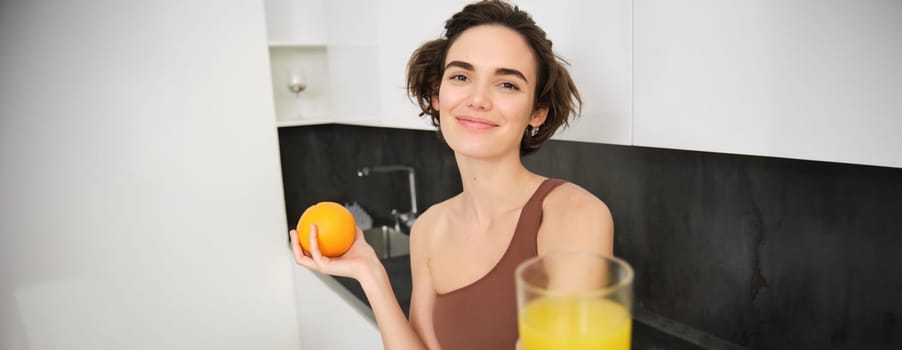 Healthy and beautiful young woman, giving you freshly squeezed glass of juice, holding an orange and smiling, taking vitamin c drink after workout, training and sport activities.