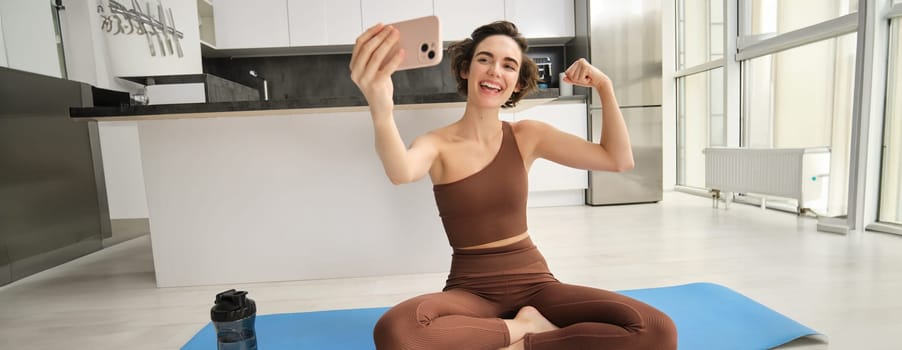 Image of young fitness girl blogger, records her workout at home, doing workout and showing muscles to telephone camera. Woman with smartphone, joining remote yoga training via mobile phone.