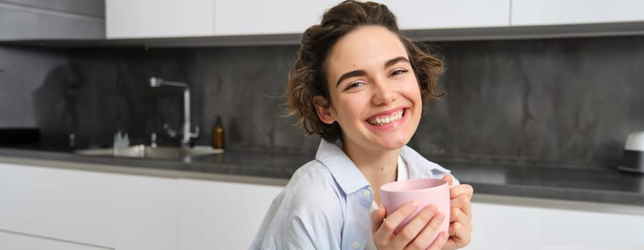Happy brunette woman drinking coffee at home. Girl with pink cup, sits in her kitchen and enjoys relaxing morning weekend.