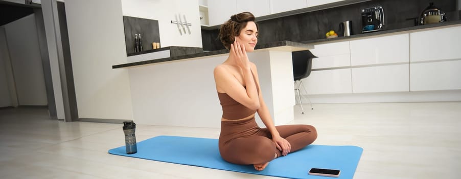 Mindfulness and healthy lifestyle. Young smiling woman sits on floor at home, practice yoga in earphones, listens to meditation app on smartphone, uses mat for exercises and workout.