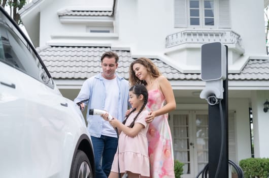 Happy little young girl learn about eco-friendly and energy sustainability as she help her family recharge electric vehicle from home EV charging station. EV car and modern family concept. Synchronos