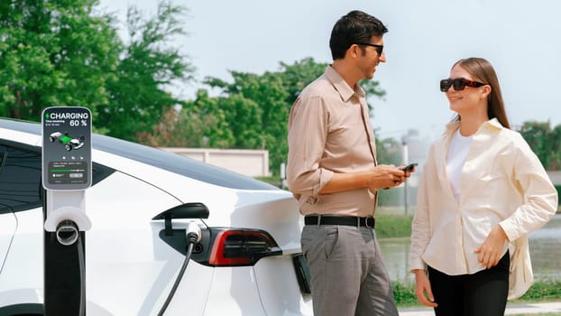 Young couple use smartphone to pay for electricity at public EV car charging station green city park. Modern environmental and sustainable urban lifestyle with EV vehicle. Expedient