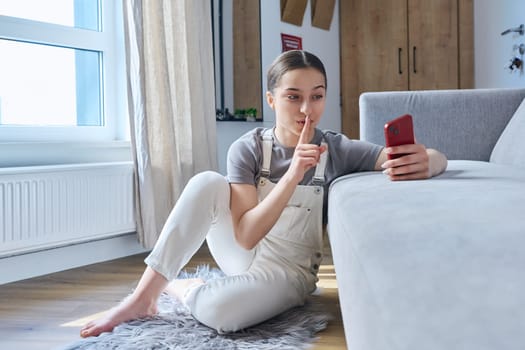 Teen girl sitting at home on floor near sofa using smartphone for video call conference, showing quiet secret sign with fingers at phone screen. Technology leisure communication lifestyle adolescence