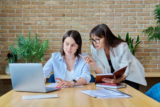 Two serious business women colleagues sitting at large table in office typing on laptop. Business ceo work law finance mentoring consulting teamwork people job concept