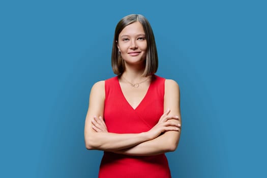 Portrait of young confident woman in red on blue studio background. Successful fashionable female with crossed arms looking at camera. Business, work, services, education, fashion beauty professions