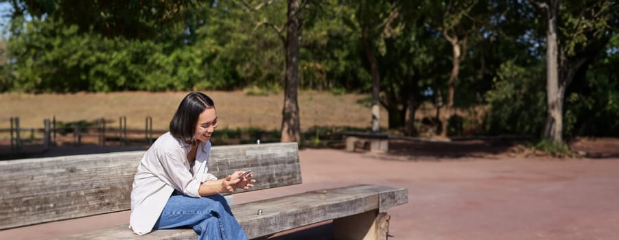 Happy smiling asian girl sitting on bench in park and browsing on social media, holding smartphone, using mobile phone app.