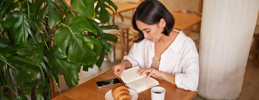 Beautiful young asian woman sitting in cafe with a book, eating croissant and reading, drinking cup of coffee. Wellbeing and self-care