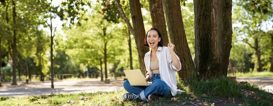 Enthusiastic young asian girl, sitting with laptop beside tree in green sunny park, celebrating, triumphing and smiling.