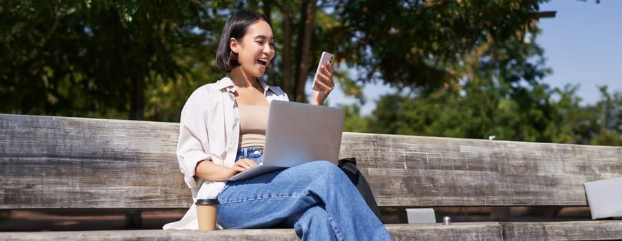 Excited young asian girl, looking at her smartphone, while sitting with laptop outdoors in sunny park.