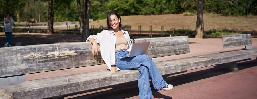 Portrait of beautiful young woman sitting on bench in park, using laptop and drinking coffee, relaxing outdoors on summer day.