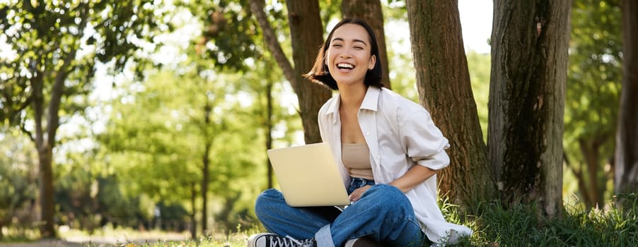 Portrait of asian girl with laptop, sitting near tree in park on sunny summer day, working on remote, e-learning with computer.