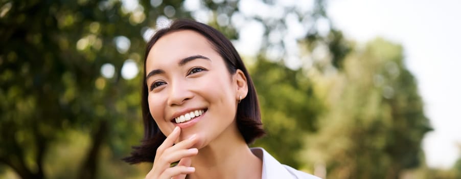 Portrait of young asian woman, laughing and smiling, looking happy while enjoying the walk in park, posing near green trees.