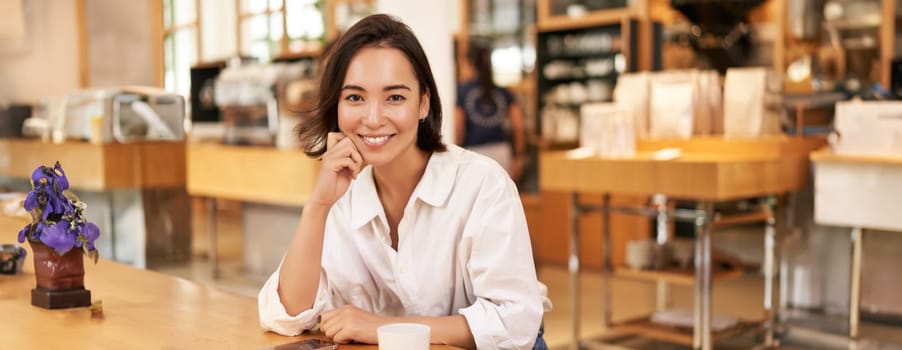 Portrait of asian businesswoman, sitting on table in cafe, drinking coffee, reading on smartphone app, smiling at camera.