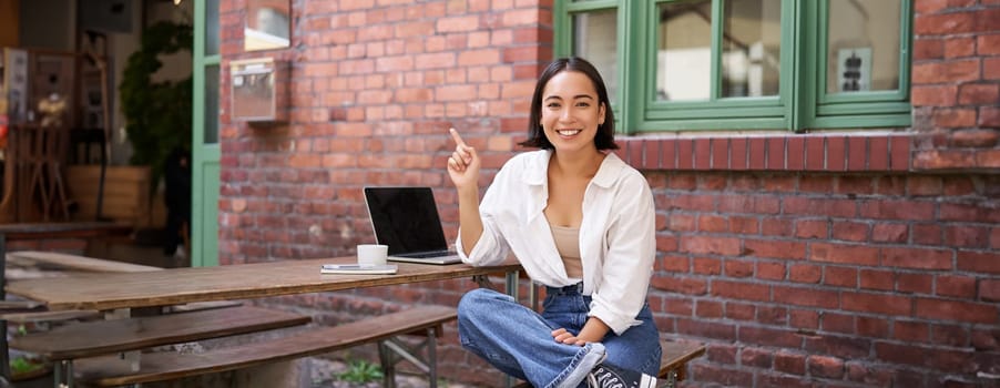 stylish modern asian girl with laptop, sitting in cafe, looking amazed and pointing at upper right corner banner, showing info advertisement.