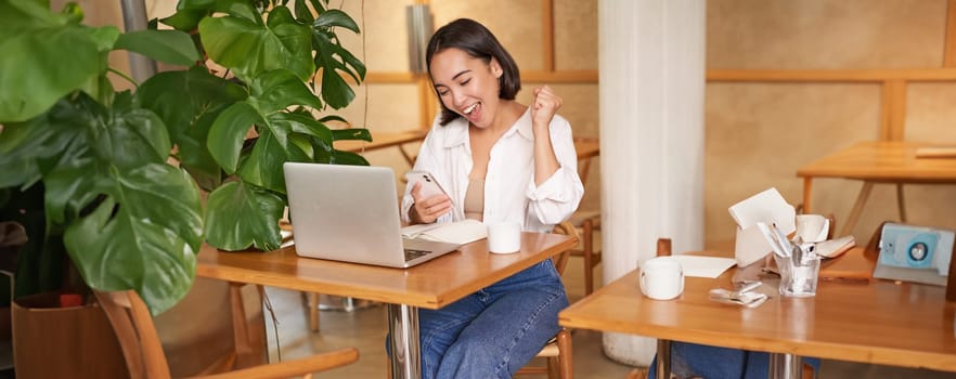 Full body shot of young asian girl sits in cafe with laptop, sees good news on smartphone and celebrates, triumphs from goal or achievement.