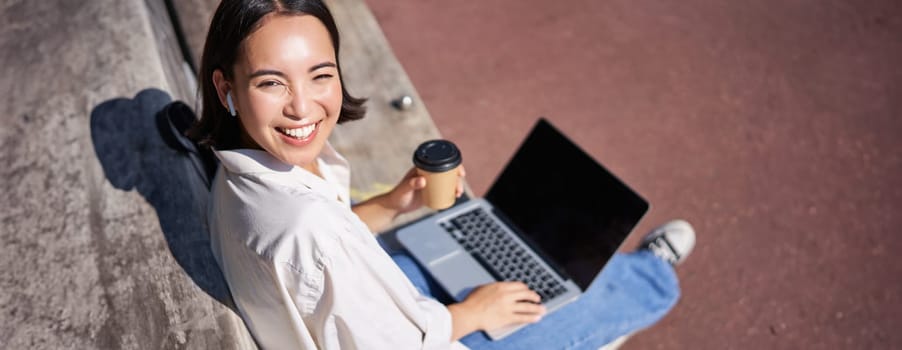 Beautiful young asian female student, sitting with laptop on bench in park, drinking takeaway coffee and listening music, working remotely, studying outdoors.