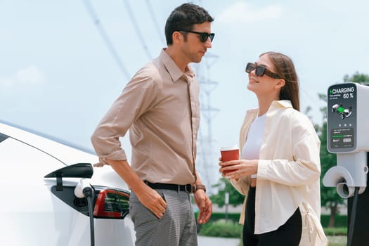 Young couple with coffee recharge EV car battery at charging station connected to electrical industrial power grid tower. Couple with shopping and travel using eco electric car lifestyle. Expedient