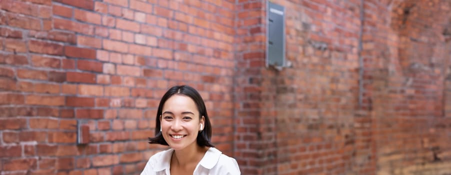 Stylish asian girl works in coworking with laptop, makes notes, uses wireless earphones and smiles, sits near brick wall in office.