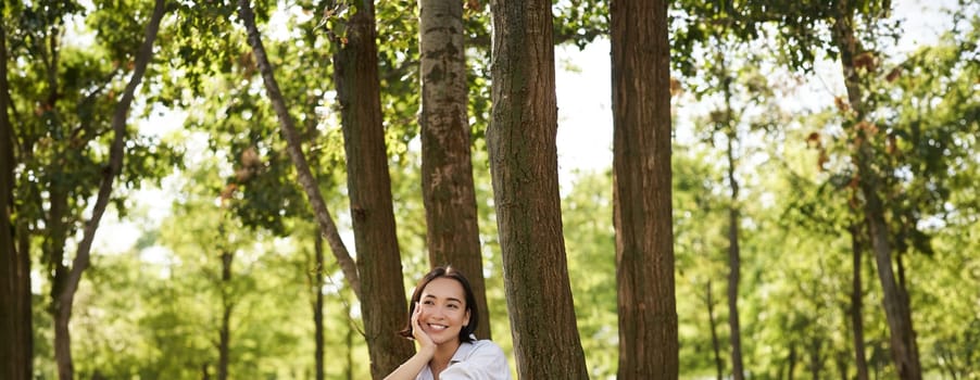 Vertical portrait of young asian woman reading her book in park, looking romantic and smiling, leaning on tree, resting outdoors.