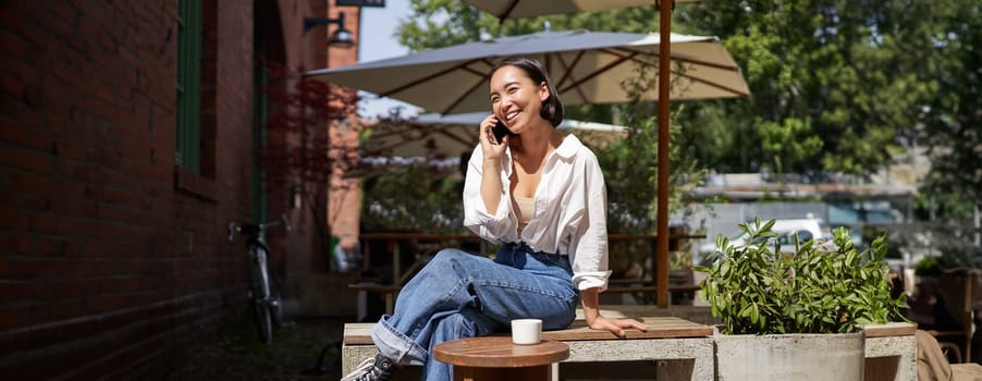 People and communication. Stylish asian girl sits in outdoor cafe with cup of coffee and smartphone, using mobile phone.