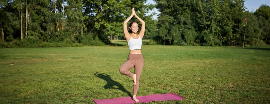 Mindfulness and wellbeing. Young woman doing yoga, standing on mat in park, making asana, meditating on fresh air, urban life concept.