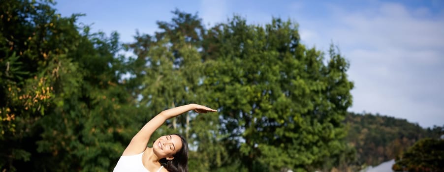 Young woman does yoga on lawn in park, stretching on fitness mat, wellbeing concept.