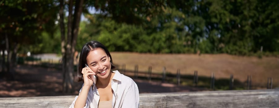 Smiling asian girl relaxing in park, talking on smartphone, having a mobile call while resting outdoors.