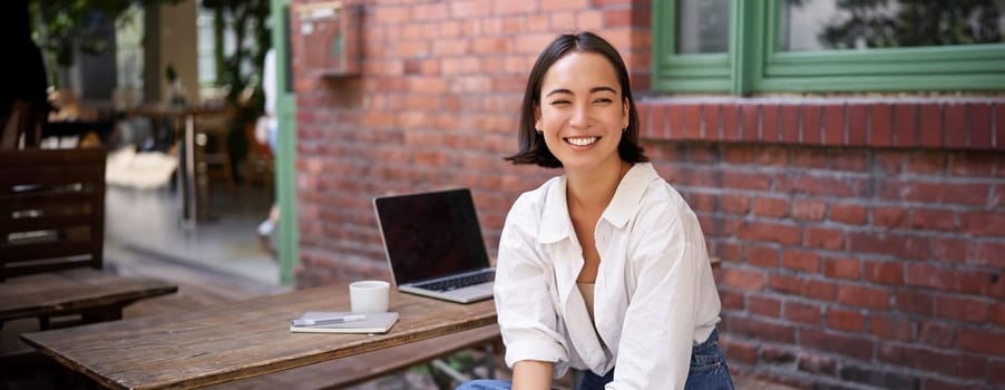 Beautiful korean girl with laptop, sitting in outdoor cafe, drinking coffee and using computer. Young people and recruitment concept