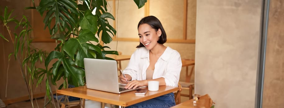 Stylish businesswoman with laptop, sitting in cafe and working on computer, managing business and drinking coffee.
