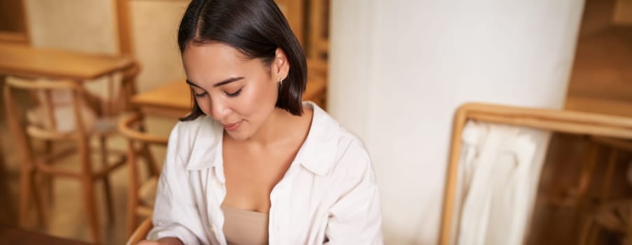 Vertical shot of young smiling woman with book, reading in cafe, enjoying morning breakfast coffee and croissant.