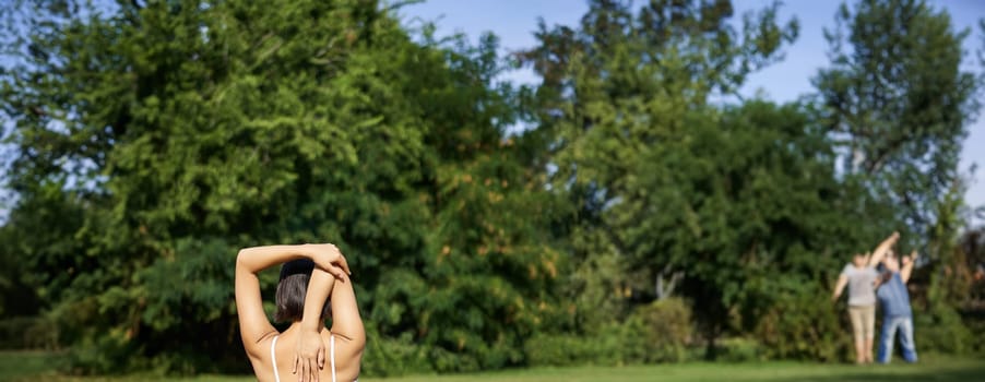 Rear view of sportswoman in middle of workout, stretching her amrs behind back, sitting on rubber yoga mat on lawn in park.