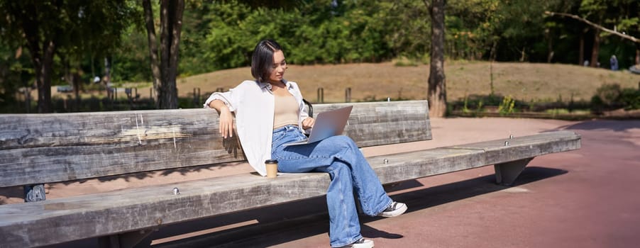 Portrait of beautiful young woman sitting on bench in park, using laptop and drinking coffee, relaxing outdoors on summer day.