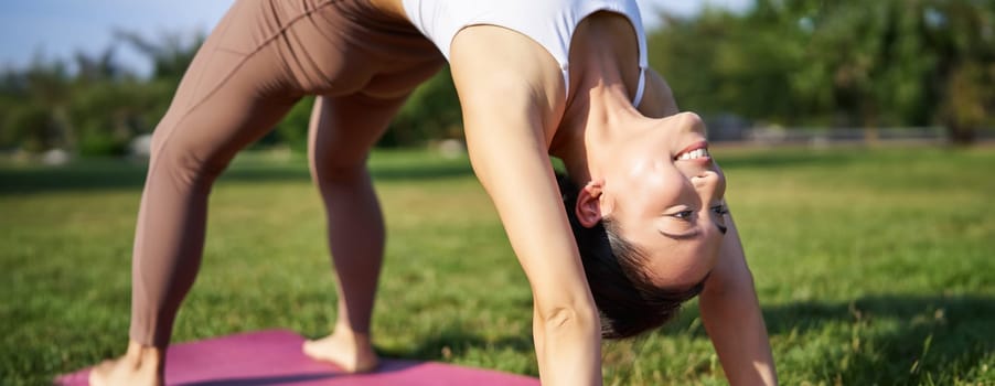 Woman smiling while making bridge asana, doing yoga in park on rubber mat.